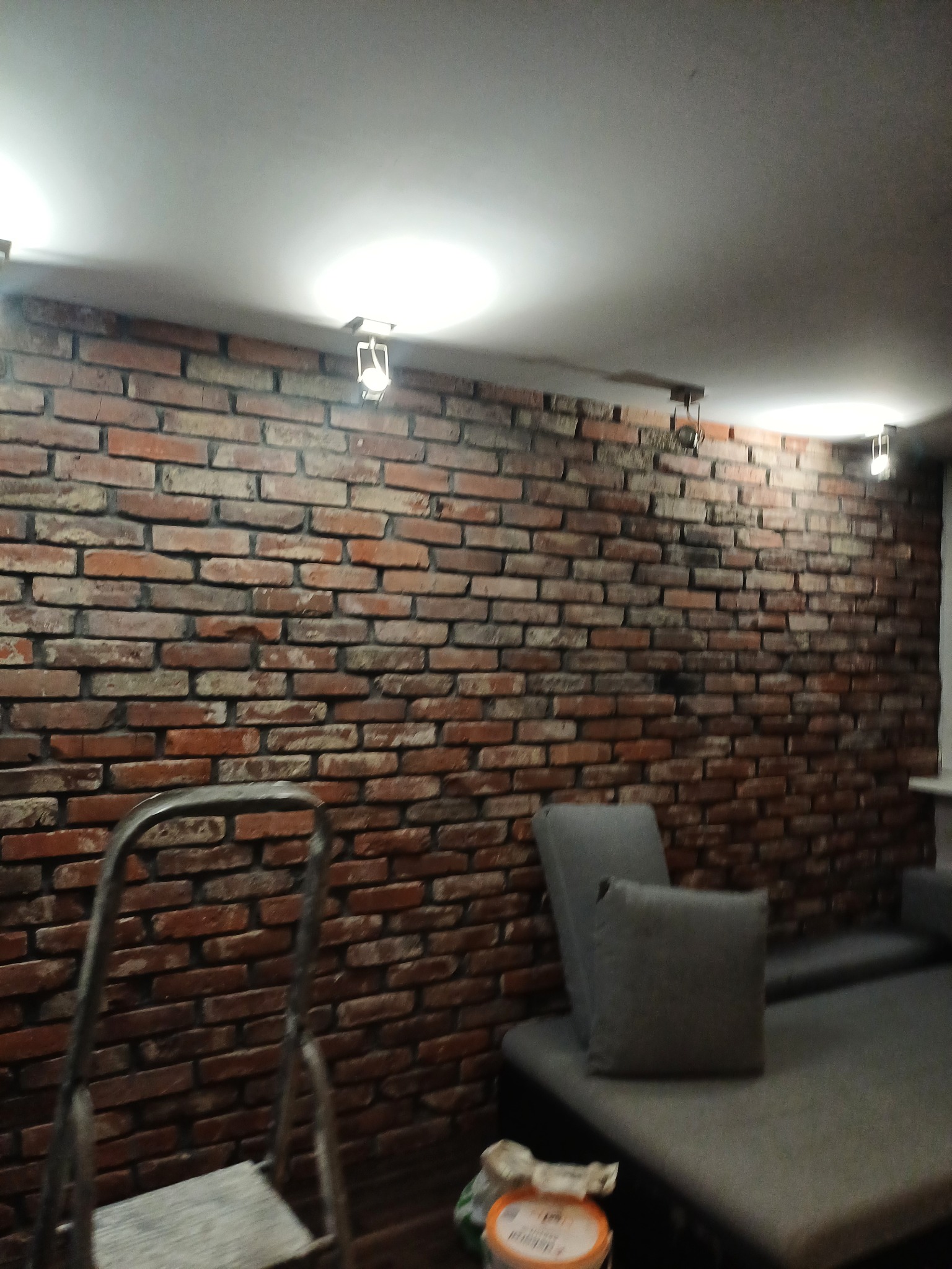 Ściana z cegły dekoracyjnej w salonie, oświetlona lampkami punktowymi, widoczna drabina malarska i fragment sofy z poduszką, na podłodze wiadro z farbą.