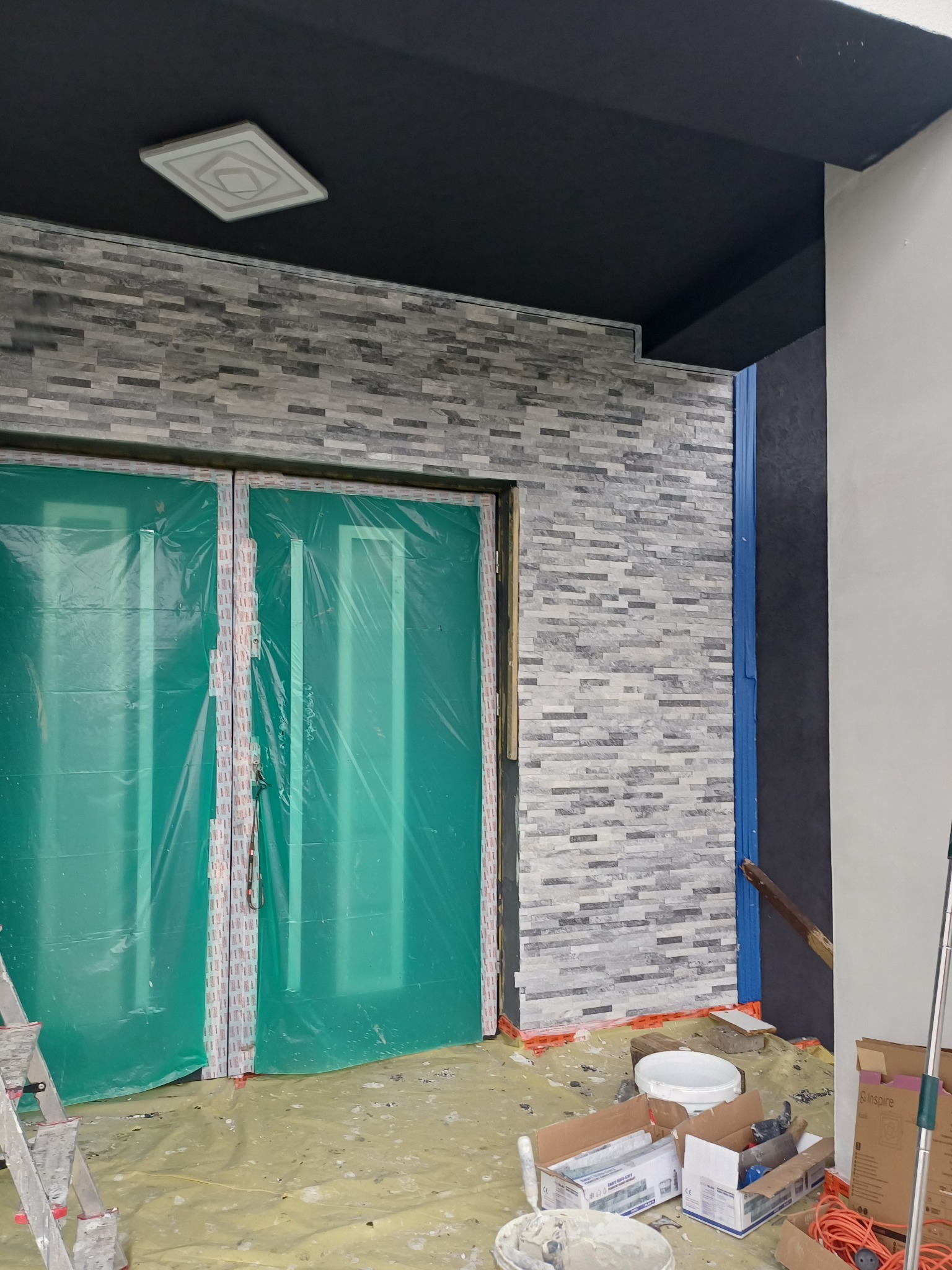 Elewacja budynku z szarej cegły, zabezpieczone folią drzwi wejściowe, widoczne narzędzia i materiały budowlane na podłożu zabezpieczonym folią malarską.