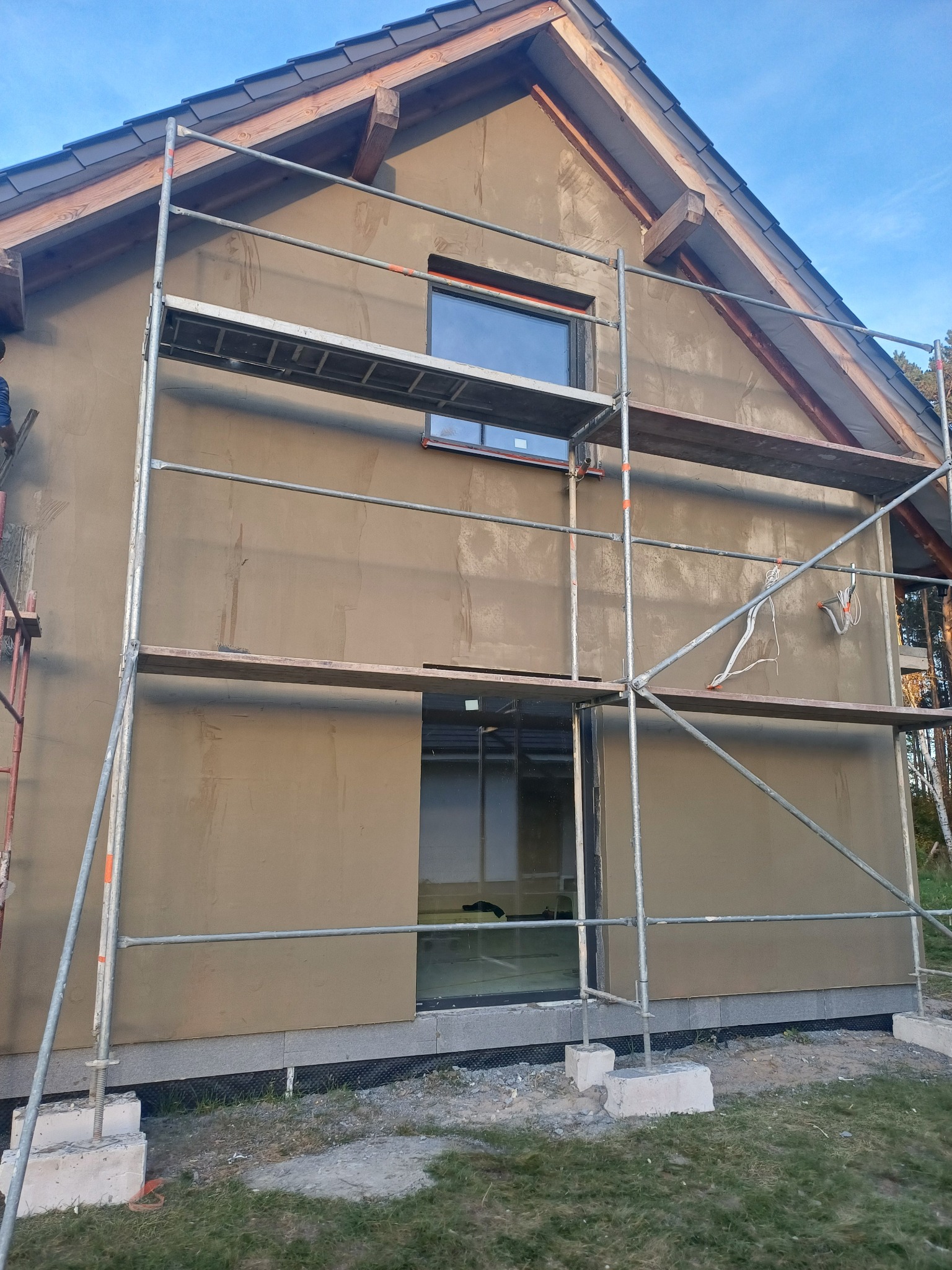 Dom w trakcie nakładania warstwy tynku zewnętrznego, widoczne rusztowanie i wystające kable elektryczne.