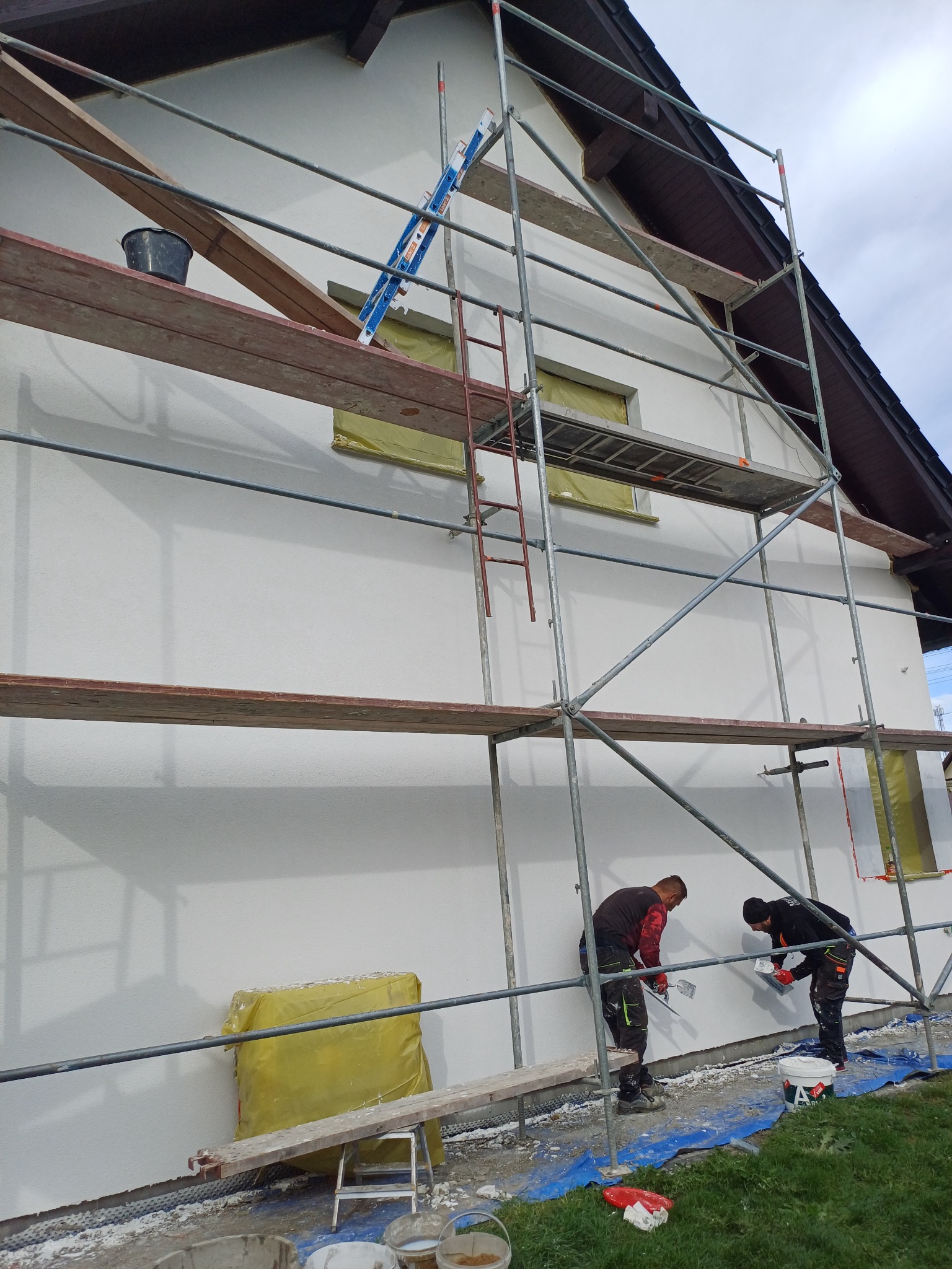 Dwóch pracowników na rusztowaniu wyrównuje świeżo nałożony tynk na elewacji domu w Głogowie, widoczne narzędzia i przygotowane okna.