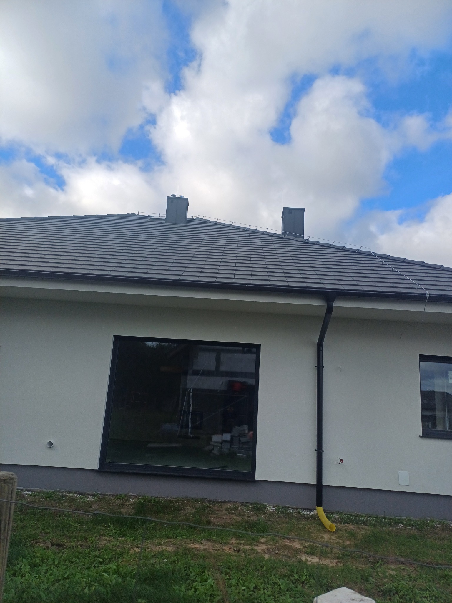 Jasna elewacja domu jednorodzinnego z grafitową dachówką, widoczne okna z czarnymi ramami, rynna z żółtym zakończeniem i fragment trawnika.