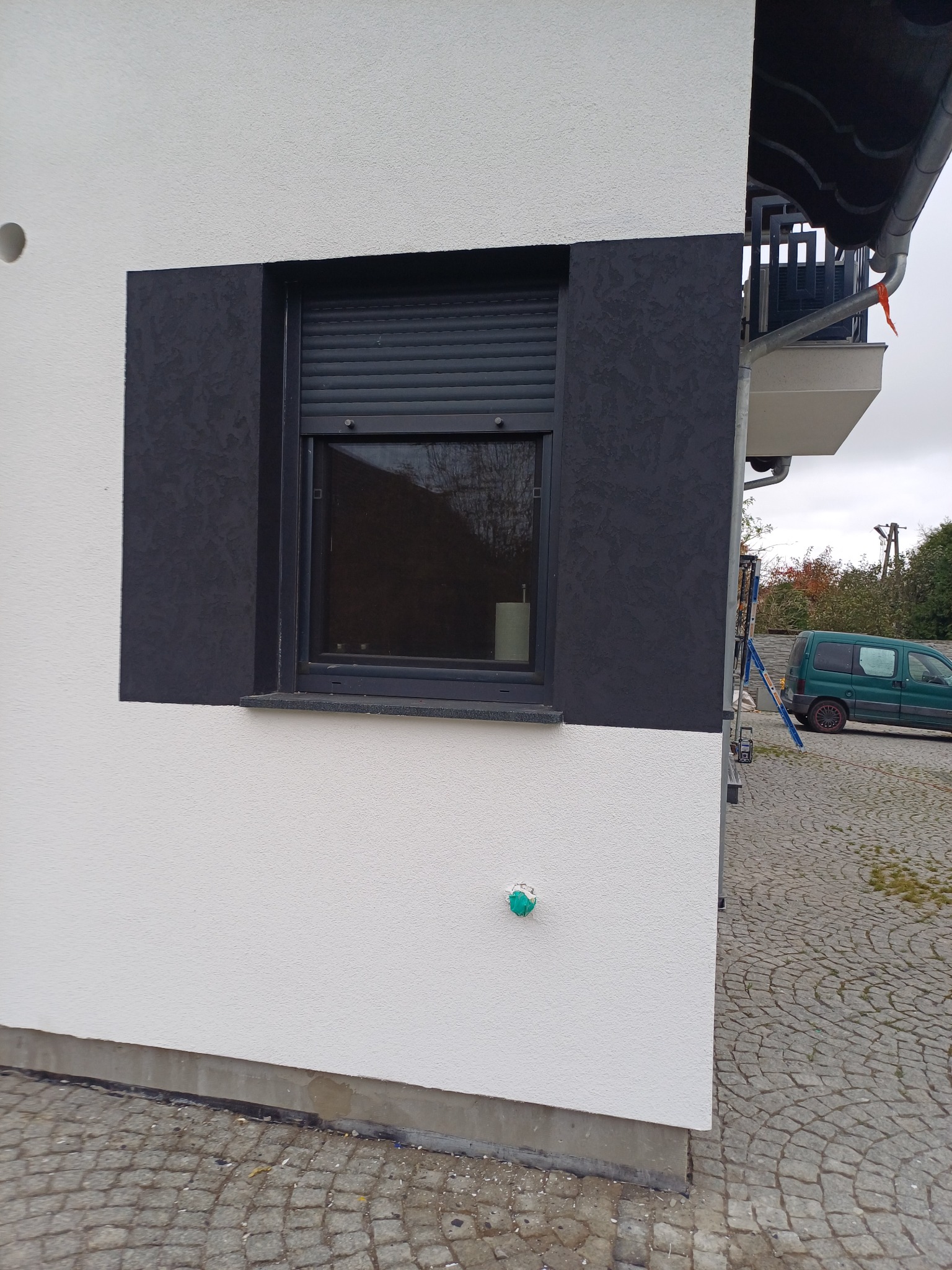 Fragment elewacji budynku z oknem z roletami, część ściany pomalowana na biało, a fragment wokół okna na ciemny grafit z wyraźną fakturą, widoczna drabina i samochód w tle.