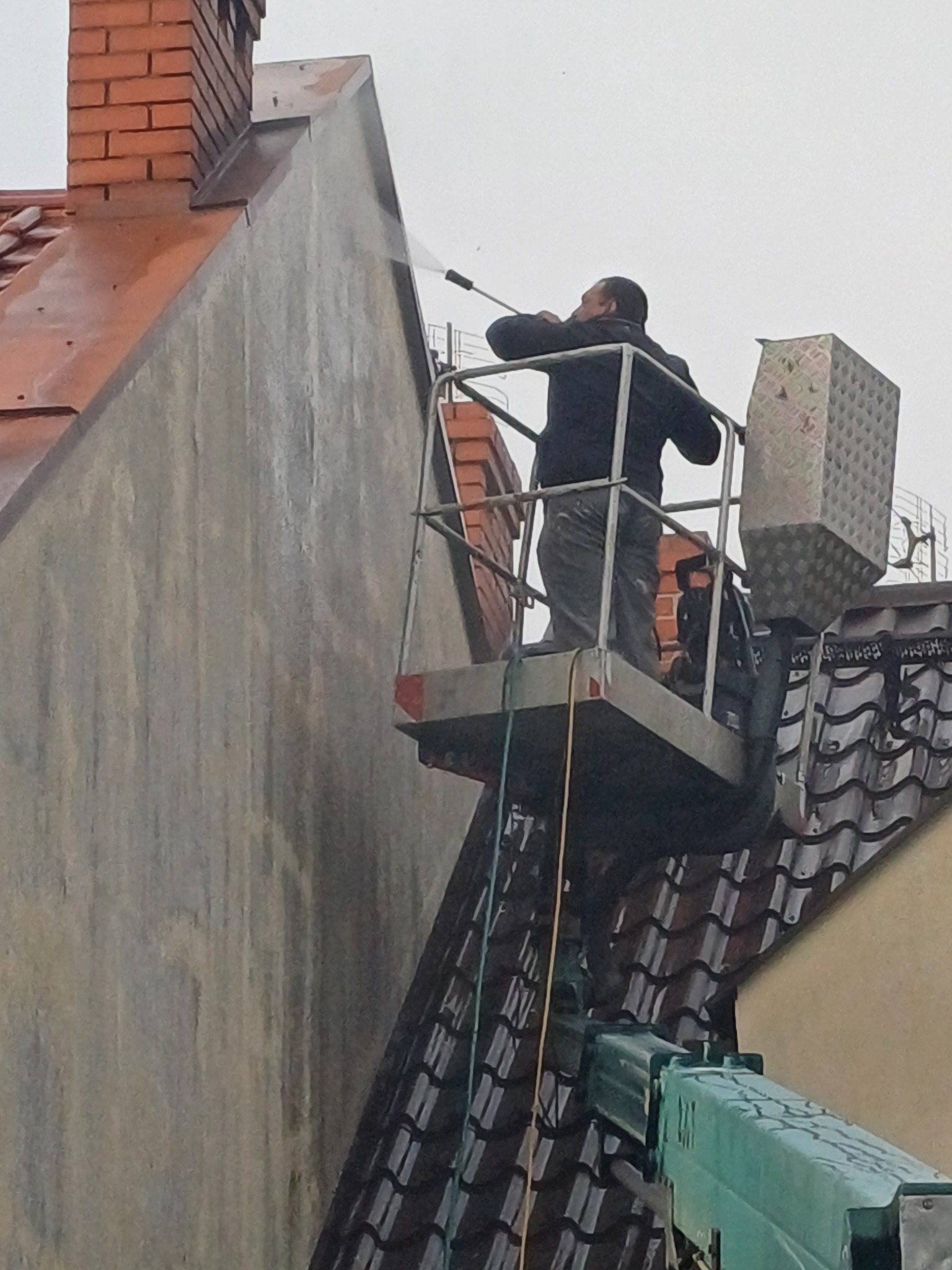 Pracownik na podnośniku koszowym czyści elewację budynku za pomocą myjki ciśnieniowej, w tle widoczny komin z czerwonej cegły i dach pokryty dachówką.