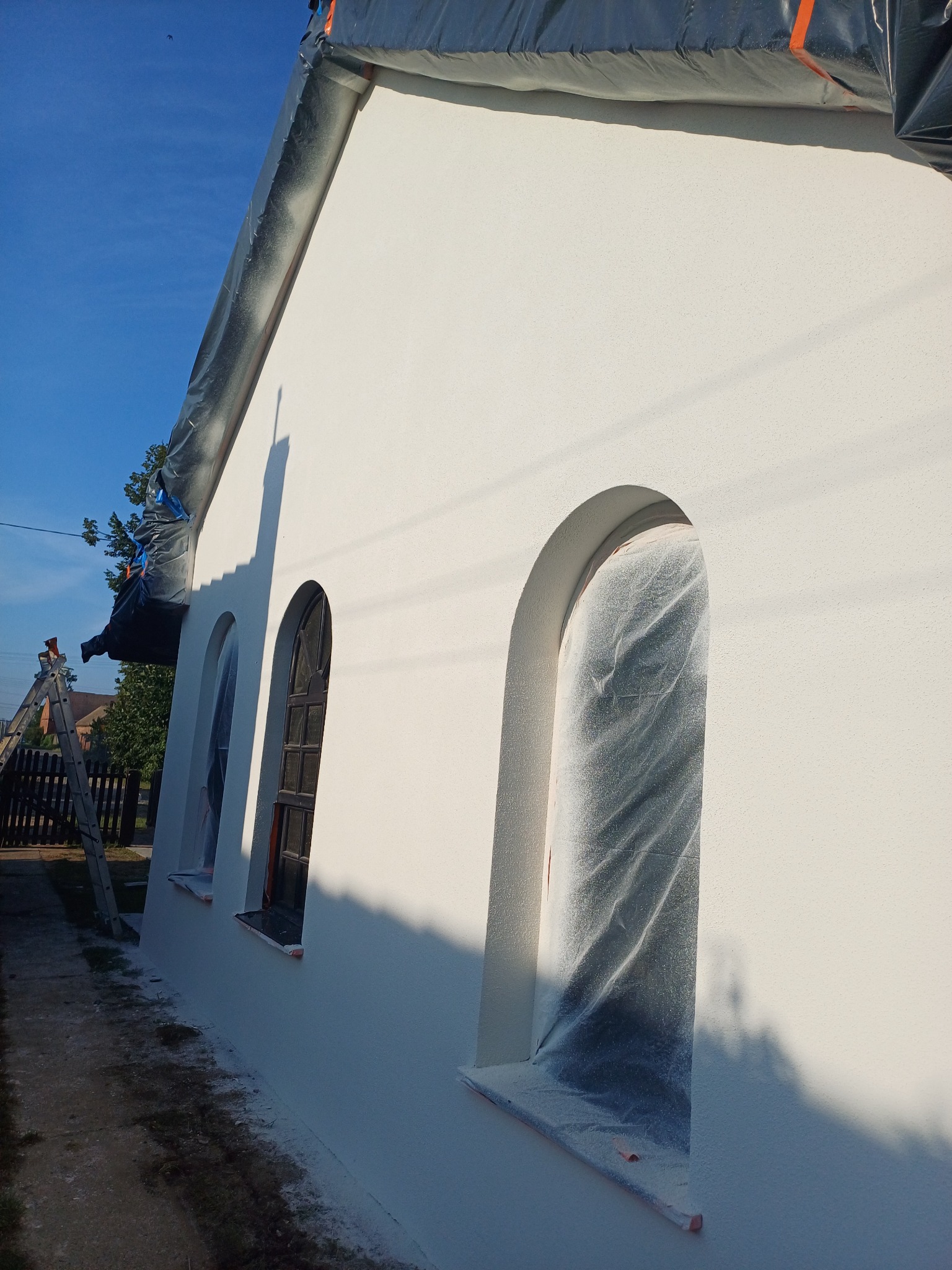 Boczny widok budynku z nową, białą elewacją, z oknami zabezpieczonymi folią, drabina oparta o ścianę, widoczne ślady świeżo nałożonego tynku na chodniku.