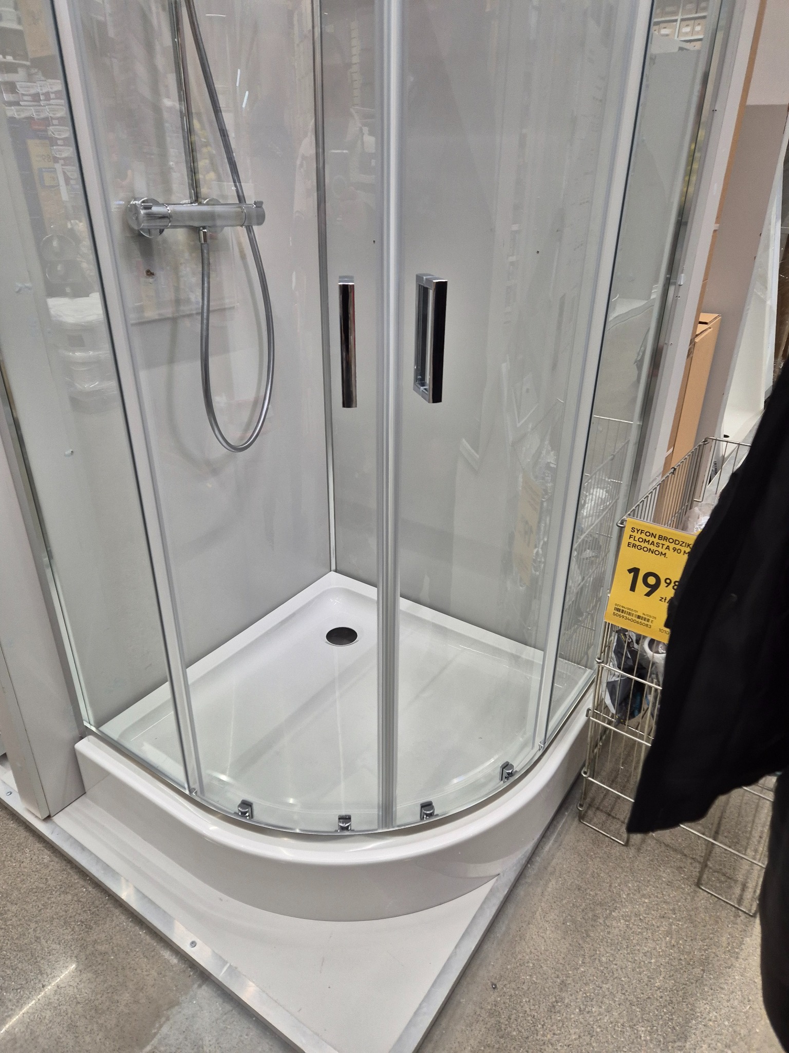 Kabiną prysznicowa z brodzikiem, widoczny chromowany panel sterujący i szklane drzwi z metalowymi uchwytami, w tle regał z akcesoriami łazienkowymi.