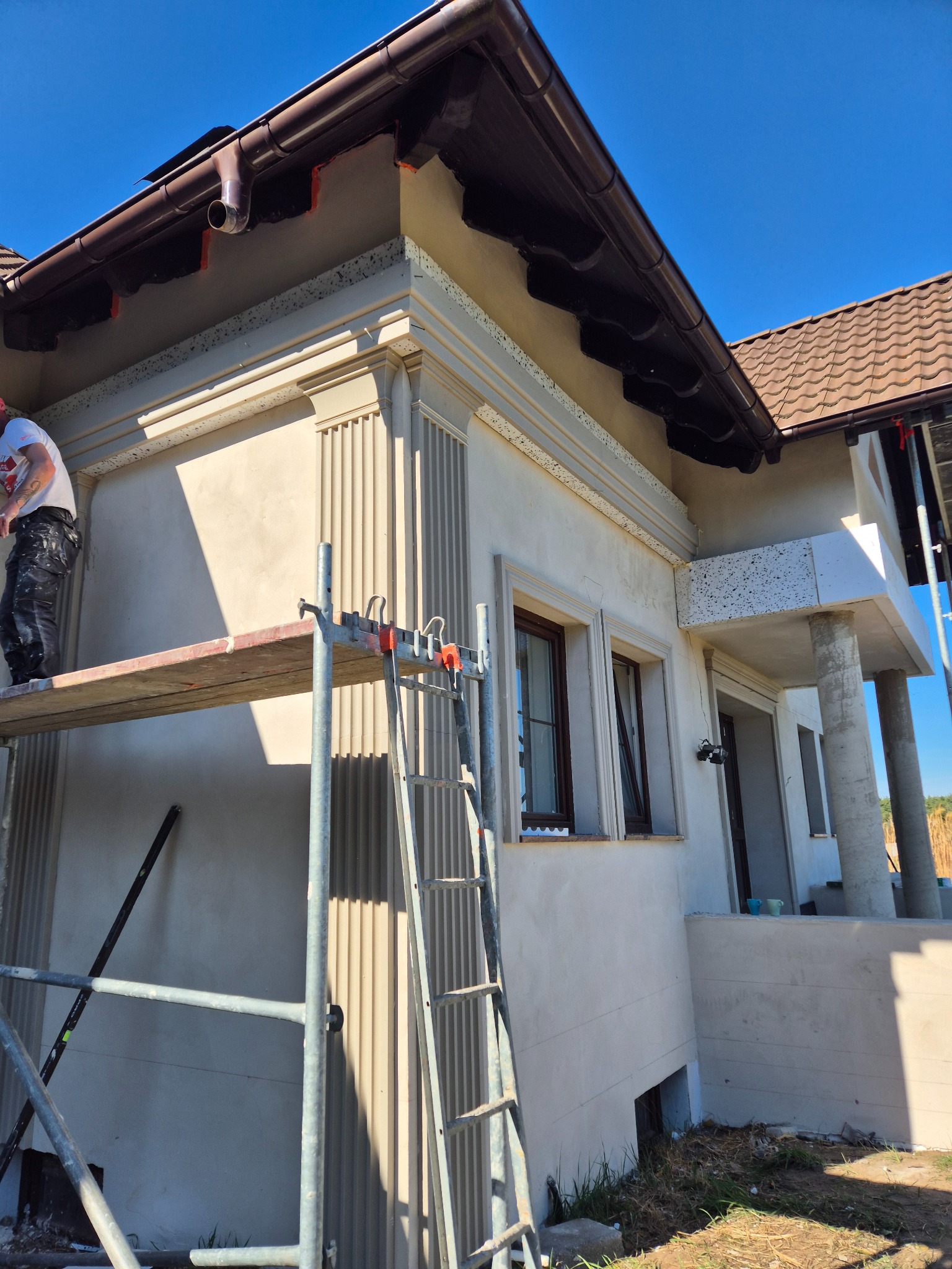 Elewacja budynku z dekoracyjnymi elementami architektonicznymi w trakcie prac wykończeniowych, widoczny pracownik na rusztowaniu, brązowe okna i dachówka.