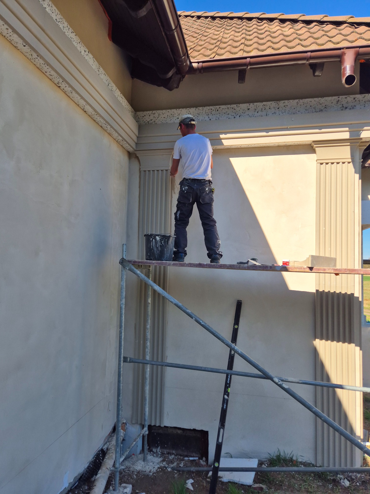 Pracownik na rusztowaniu, w trakcie nakładania zaprawy na elewację budynku z dekoracyjnymi elementami architektonicznymi, widoczny kubeł z zaprawą i poziomica.