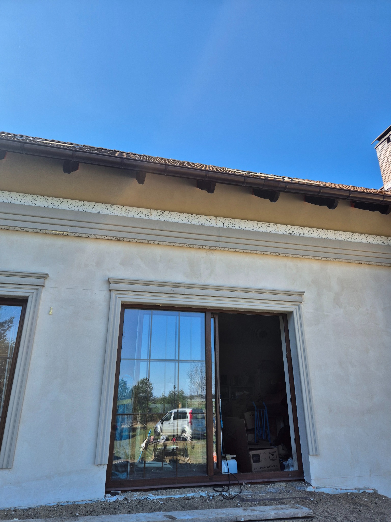 Elewacja budynku w trakcie prac wykończeniowych, widoczne okna z brązowymi ramami, fragment dachu z brązową dachówką oraz ściana pokryta jasnym tynkiem z ozdobnymi listwami.