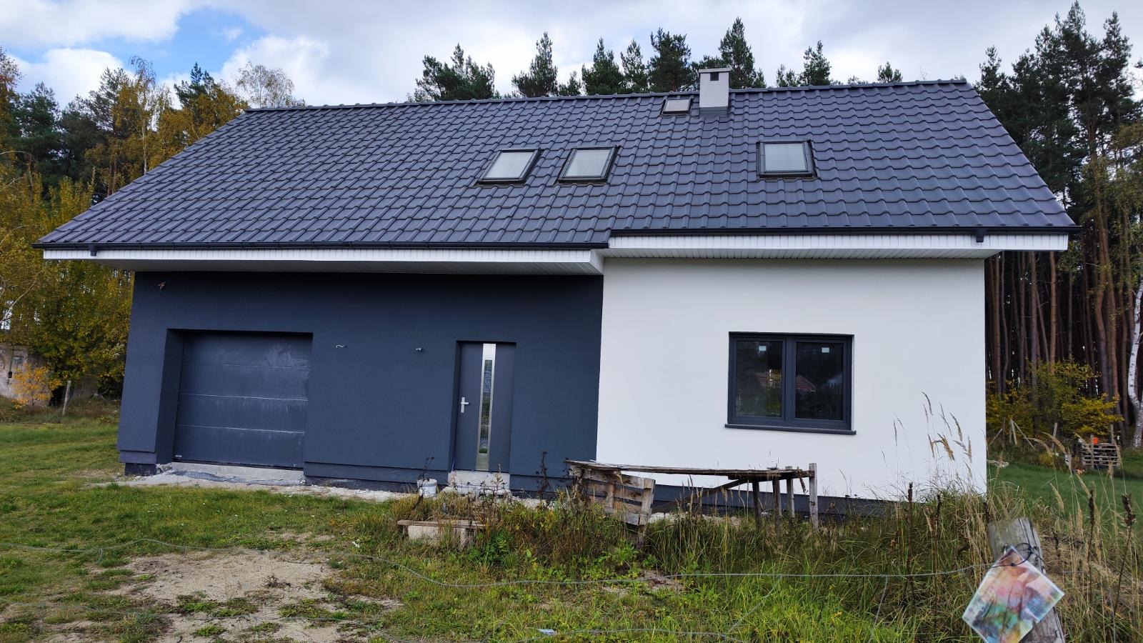 Dwukolorowy dom z ciemnoszarą elewacją garażu i wejściową oraz białą częścią z oknem. Dach z trzema oknami dachowymi. Widok z zewnątrz, trawa i drzewa w tle.