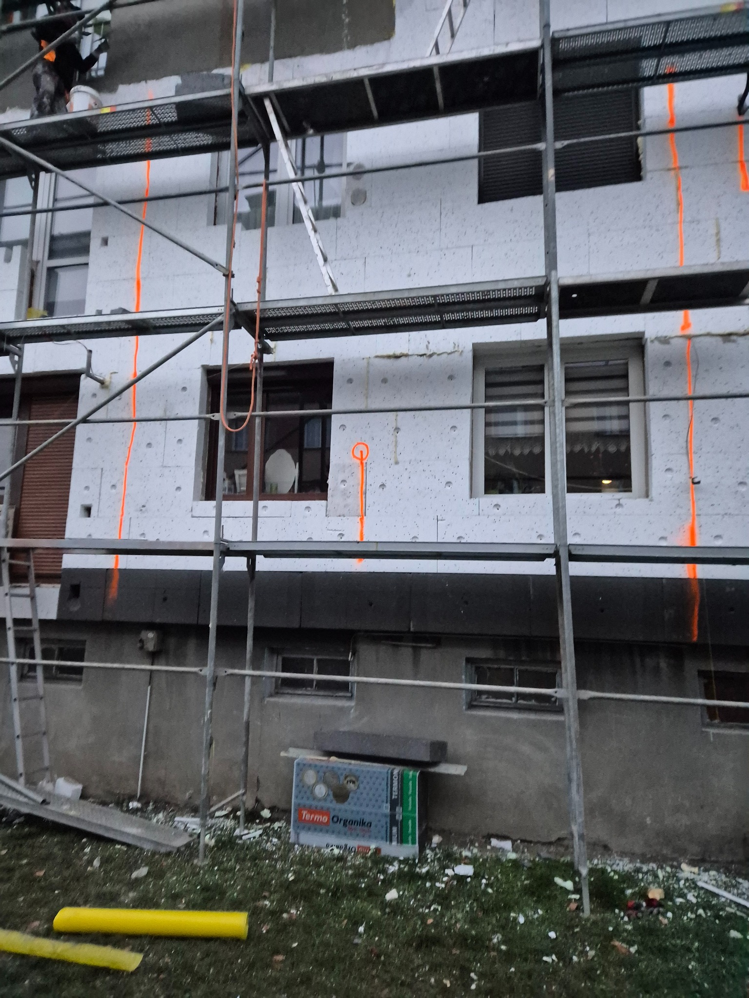 Budynek w trakcie ocieplania, widoczne rusztowania, styropian z pomarańczowymi oznaczeniami i opakowanie styropianu Terma Organika u podnóża ściany.