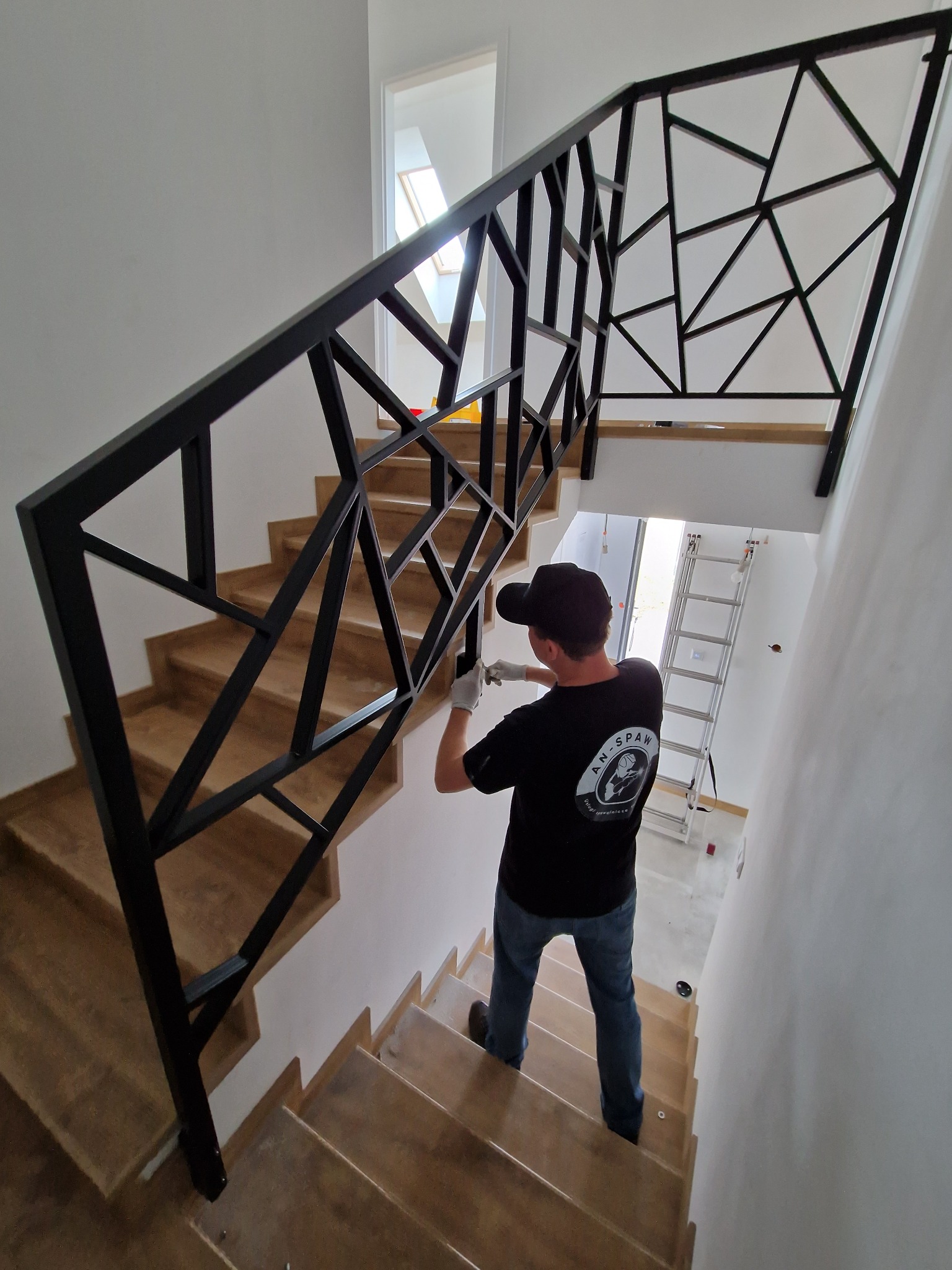 Montaż czarnej, geometrycznej balustrady metalowej na drewnianych schodach w nowoczesnym wnętrzu; pracownik w czarnej koszulce z logo firmy AN-SPAW wykonuje prace wykończeniowe.