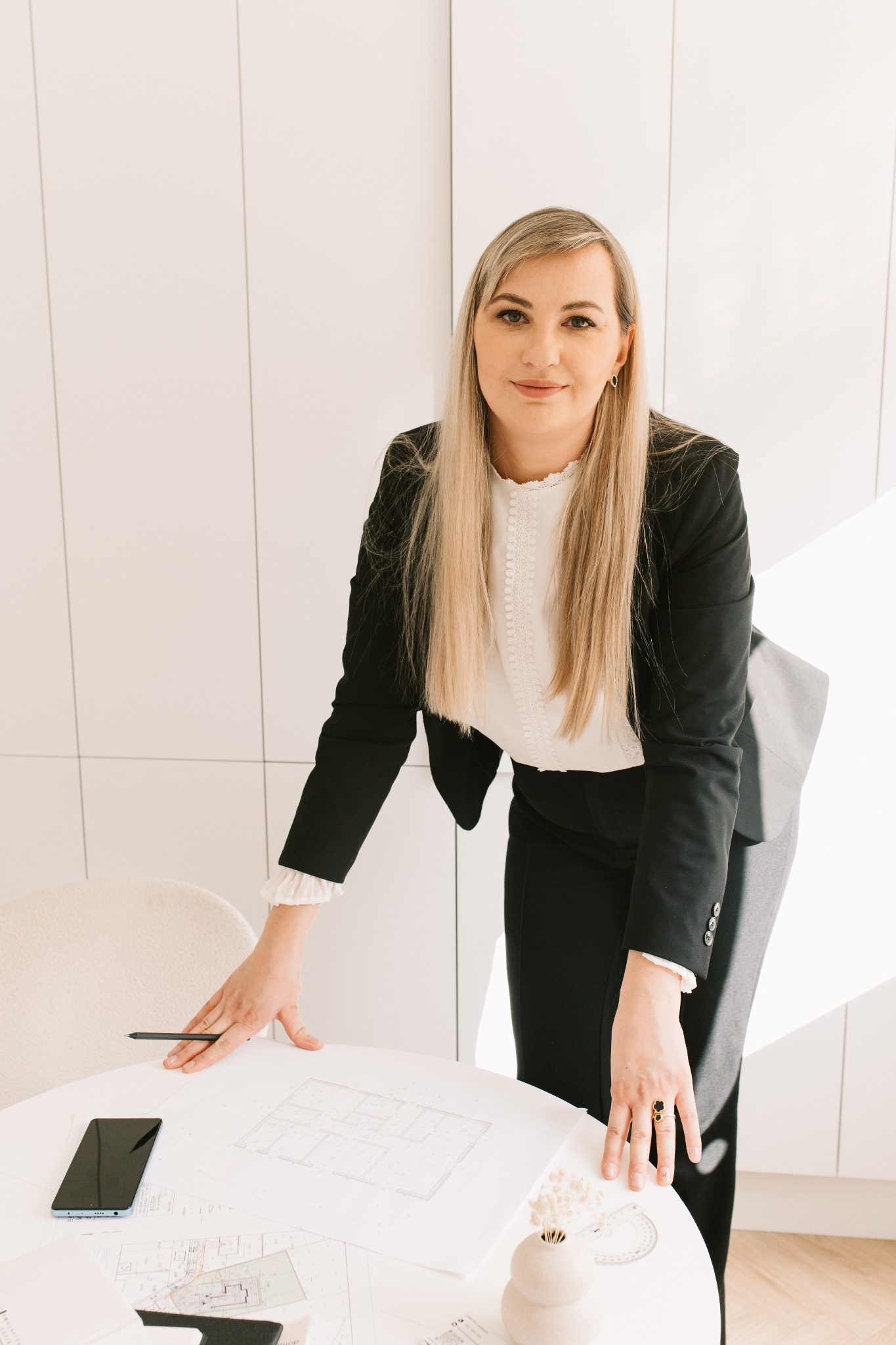 Kobieta w czarnym żakiecie pochyla się nad okrągłym stołem z planami architektonicznymi, telefonem i przyborami. Jasne wnętrze, minimalistyczny styl.