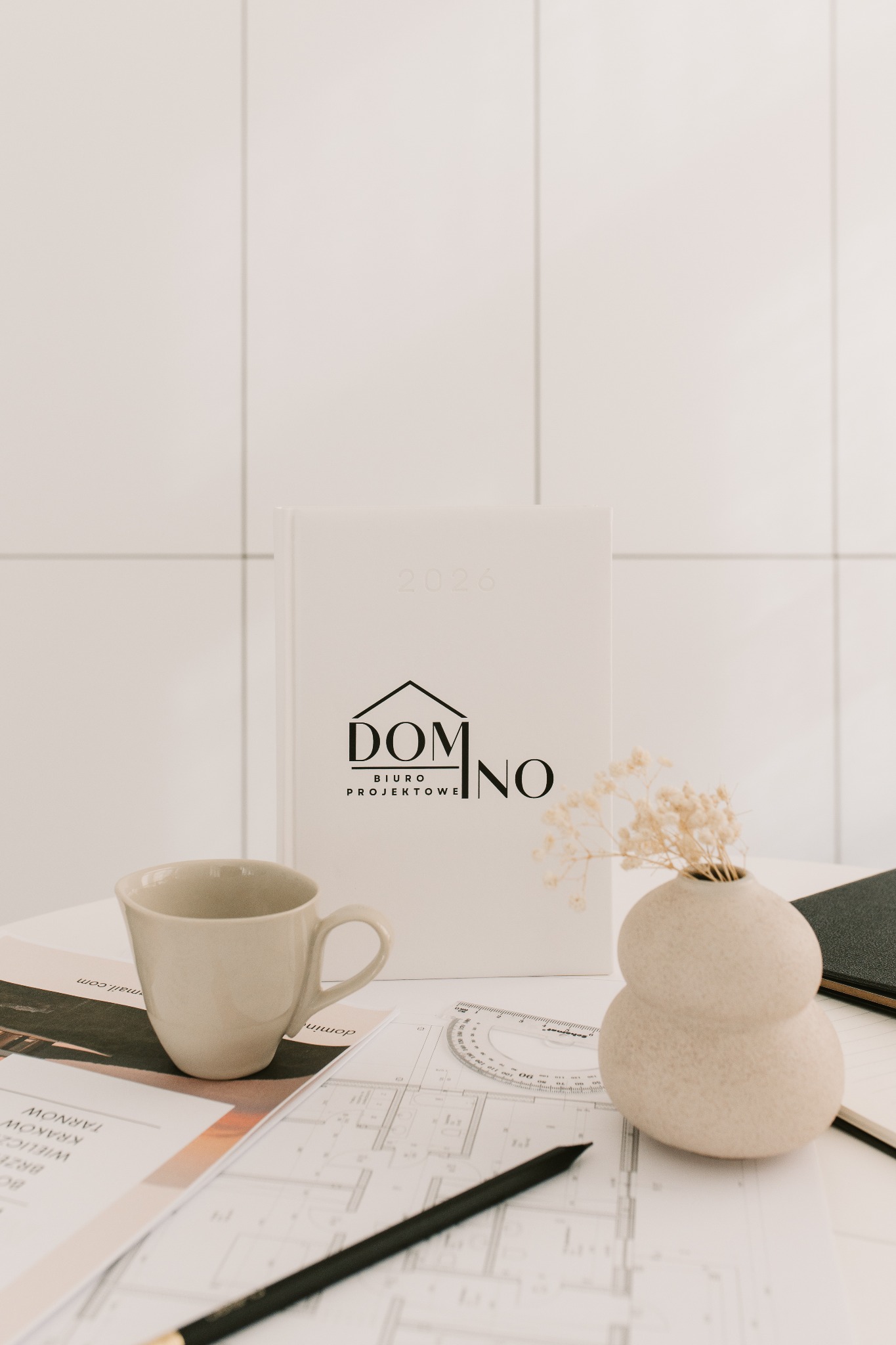 Biuro projektowe: Plan architektoniczny z kubkiem, ołówkiem, wazonem z suszem i kalendarzem na tle białych płytek. Minimalistyczny design, stonowana kolorystyka.