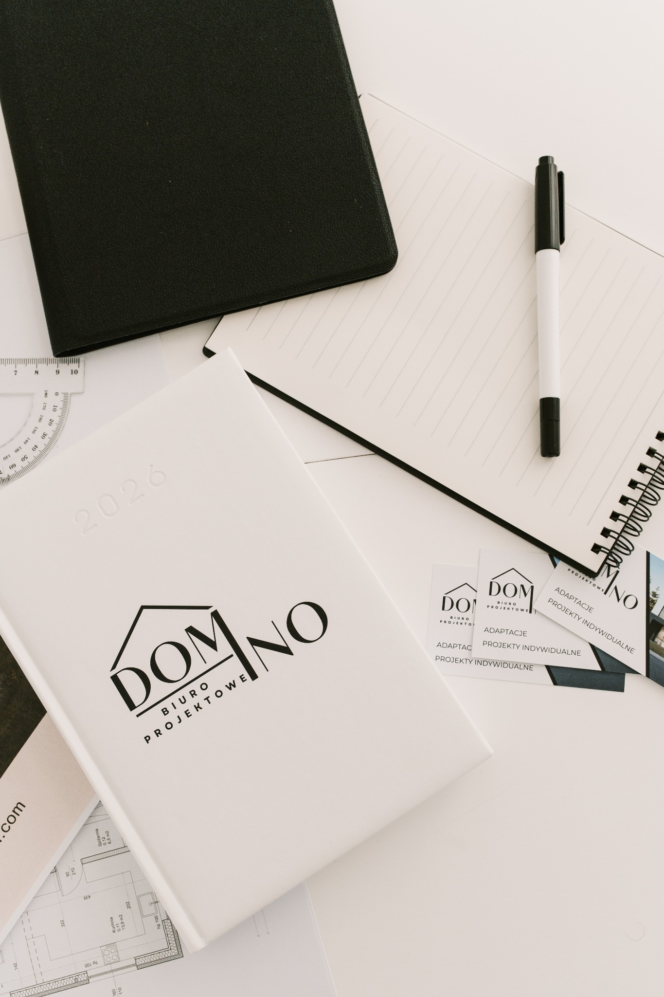 Biały notatnik z logo biura projektowego 'Domino' na tle planów architektonicznych, wizytówek i czarnego notesu. Minimalistyczna kompozycja na jasnym tle.