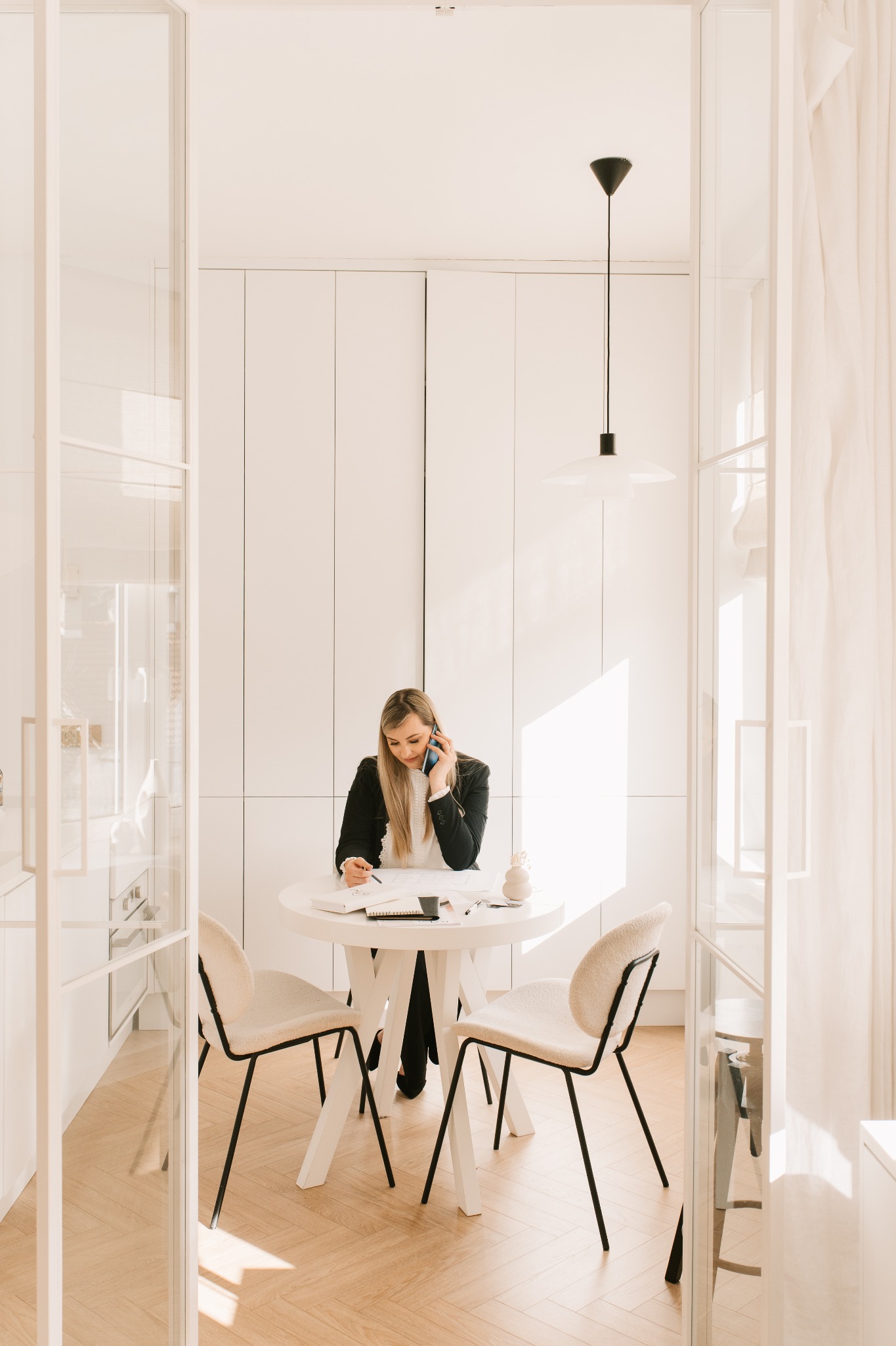 Kobieta w czarnej marynarce rozmawia przez telefon i notuje przy okrągłym, białym stole w jasnym wnętrzu. Widoczne szklane drzwi i minimalistyczna lampa.