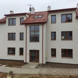 Trzykondygnacyjny budynek mieszkalny z jasną elewacją, brązową stolarką okienną i czerwoną dachówką, widziany z poziomu chodnika w pochmurny dzień.