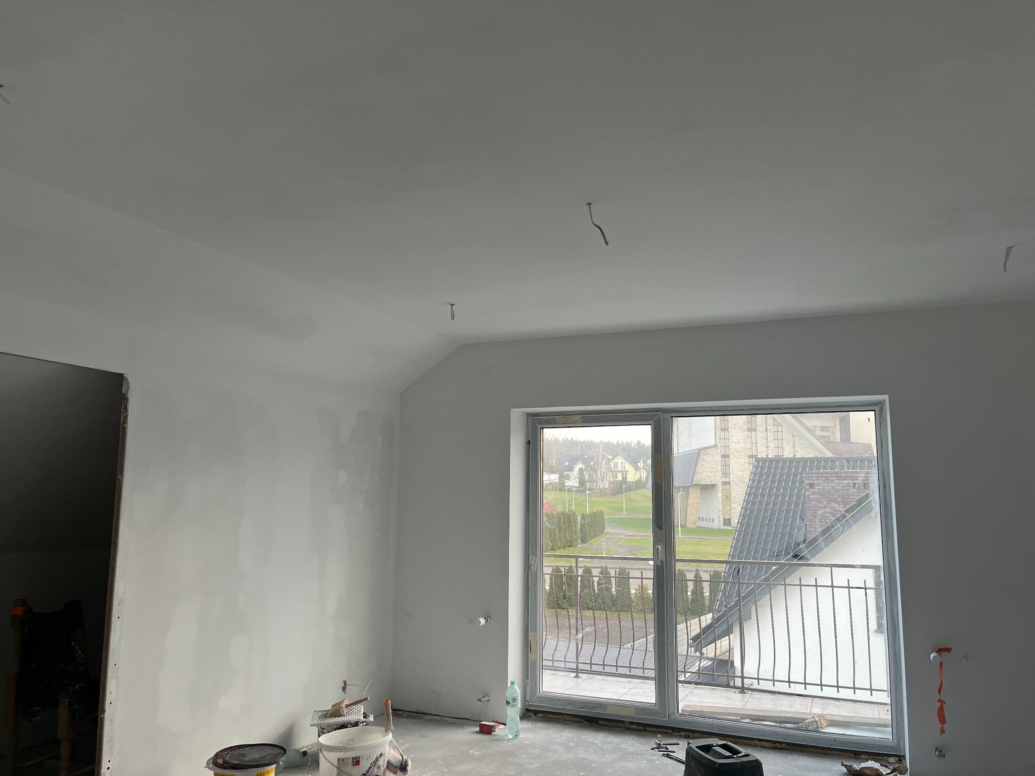 Wnętrze pokoju na poddaszu w trakcie prac wykończeniowych, ściany częściowo pokryte gładzią, widoczne narzędzia malarskie i wiadro z masą szpachlową na podłodze, okno balkonowe z widokiem na domy...
