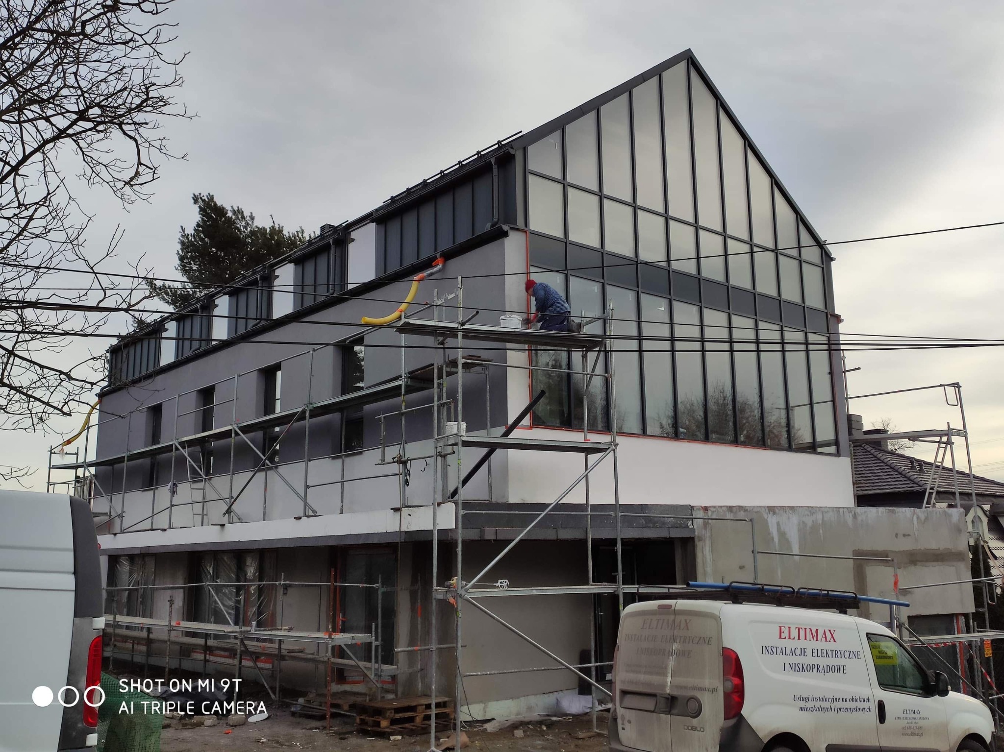 Pracownik na rusztowaniu maluje elewację nowoczesnego, przeszklonego domu w trakcie budowy, z widocznym samochodem dostawczym firmy elektrycznej w tle.