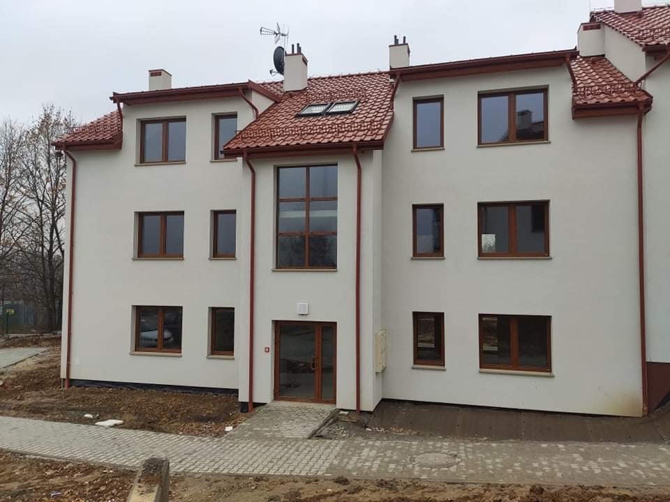 Trzykondygnacyjny budynek mieszkalny z jasną elewacją, brązową stolarką okienną i czerwoną dachówką, widziany z poziomu chodnika w pochmurny dzień.