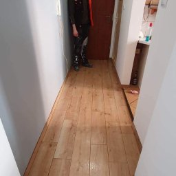 Erwin Chamera - Korytarz po remoncie z nową drewnopodobną podłogą z płytek, świeżo pomalowane ściany w odcieniach szarości i bieli, w tle widoczny pracownik w ubrudzonych farbą spodniach.