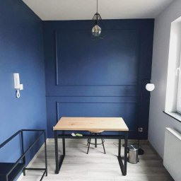 Erwin Chamera - Pomieszczenie po malowaniu na granatowo z dekoracyjnymi listwami ściennymi, drewniany blat biurka na metalowych nogach i lampa wisząca z żarówką w metalowej klatce.