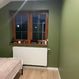 Pokój w odcieniach zieleni z drewnianym oknem, białym grzejnikiem i jasną podłogą z paneli, częściowo widoczne łóżko.