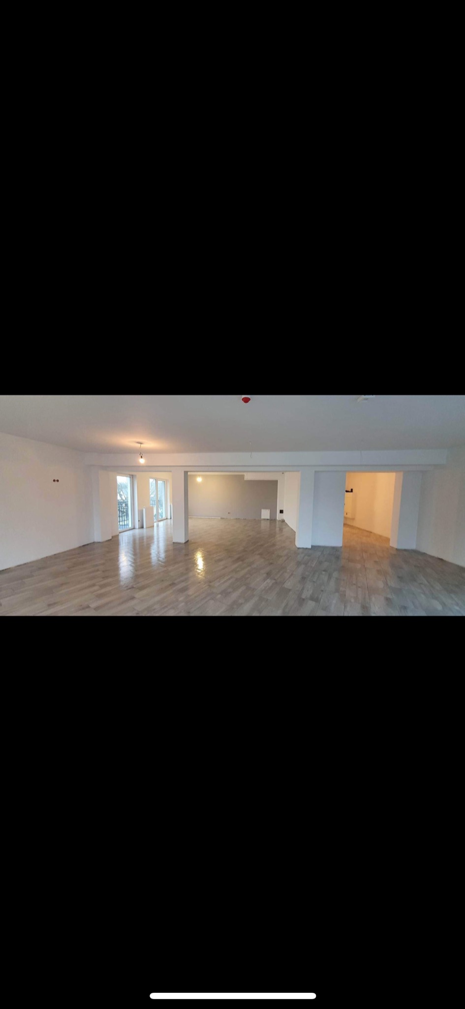 Przestronne, jasne wnętrze po remoncie, z drewnopodobną podłogą, białymi ścianami i kolumnami, widok na balkon przez przeszklone drzwi.