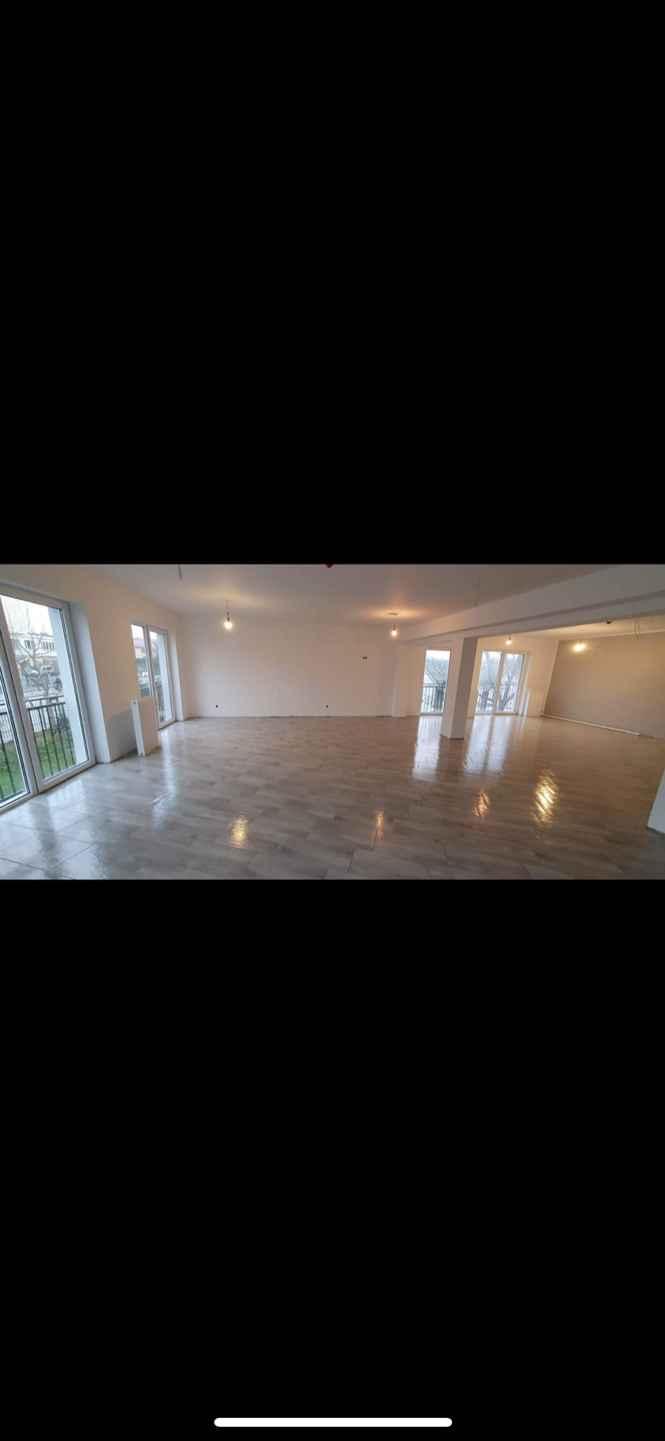 Panoramiczny widok na przestronne, jasne wnętrze z nową, błyszczącą podłogą z płytek imitujących drewno, świeżo pomalowanymi ścianami na biało i dużymi oknami balkonowymi wychodzącymi na zewnątrz.