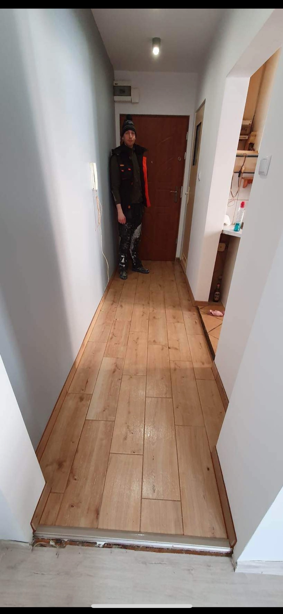Korytarz po remoncie z nową drewnopodobną podłogą z płytek, świeżo pomalowane ściany w odcieniach szarości i bieli, w tle widoczny pracownik w ubrudzonych farbą spodniach.