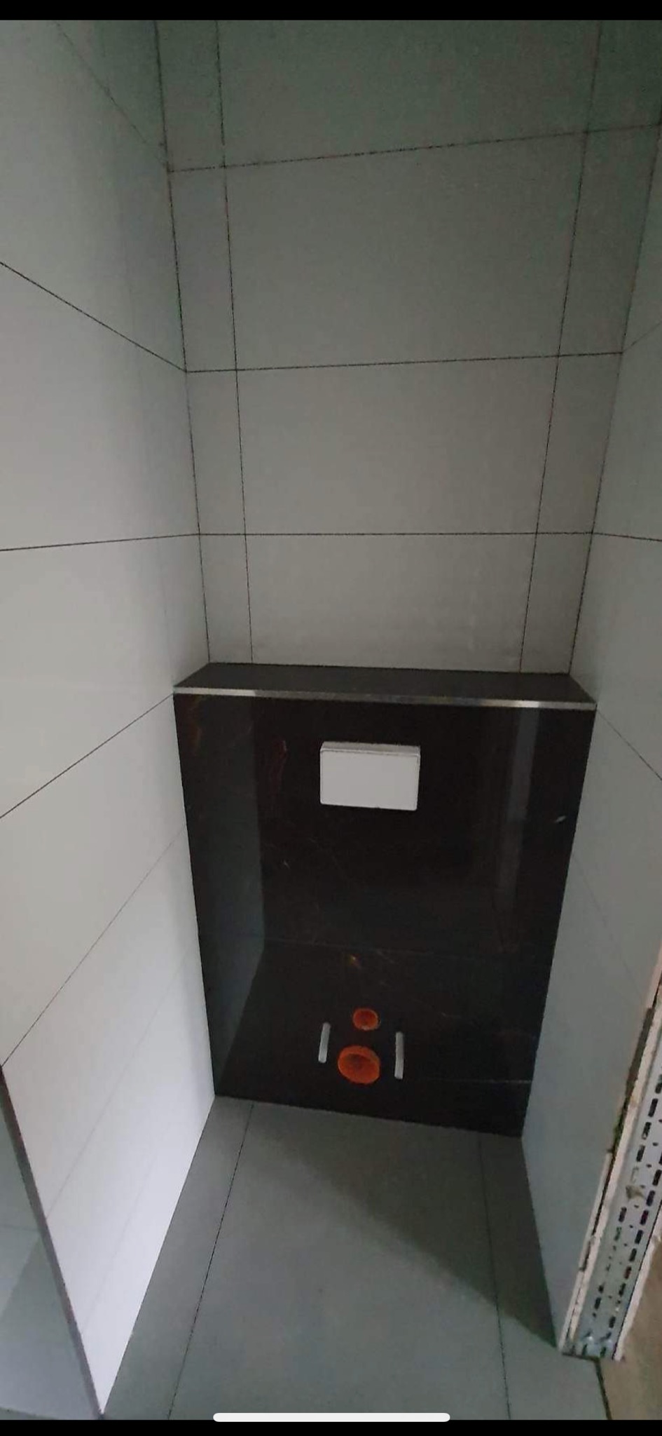 Nowoczesna, minimalistyczna łazienka w trakcie remontu: widoczna zabudowa stelaża WC z ciemnego marmuru i białym przyciskiem, białe płytki na ścianach, szare płytki na podłodze, pomarańczowe rury...