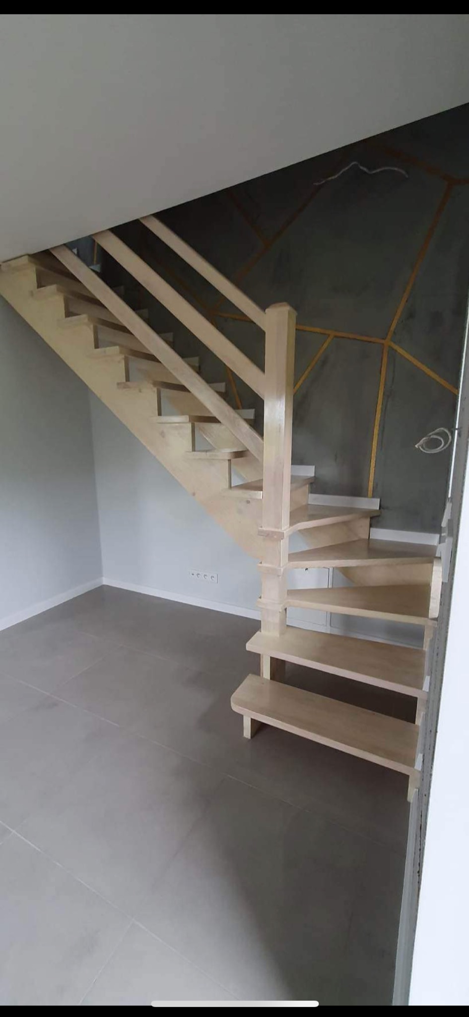 Jasne, drewniane schody z barierką, prowadzące na górę w nowym domu. Na ścianie szary tynk dekoracyjny z geometrycznym wzorem w kolorze złotym. Szare płytki na podłodze.