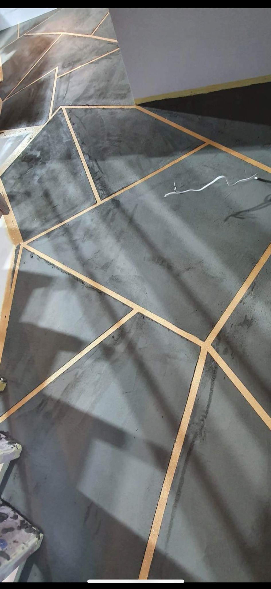 Świeżo pomalowana ściana z geometrycznym wzorem w odcieniach szarości i złota, widoczne ślady taśmy malarskiej.