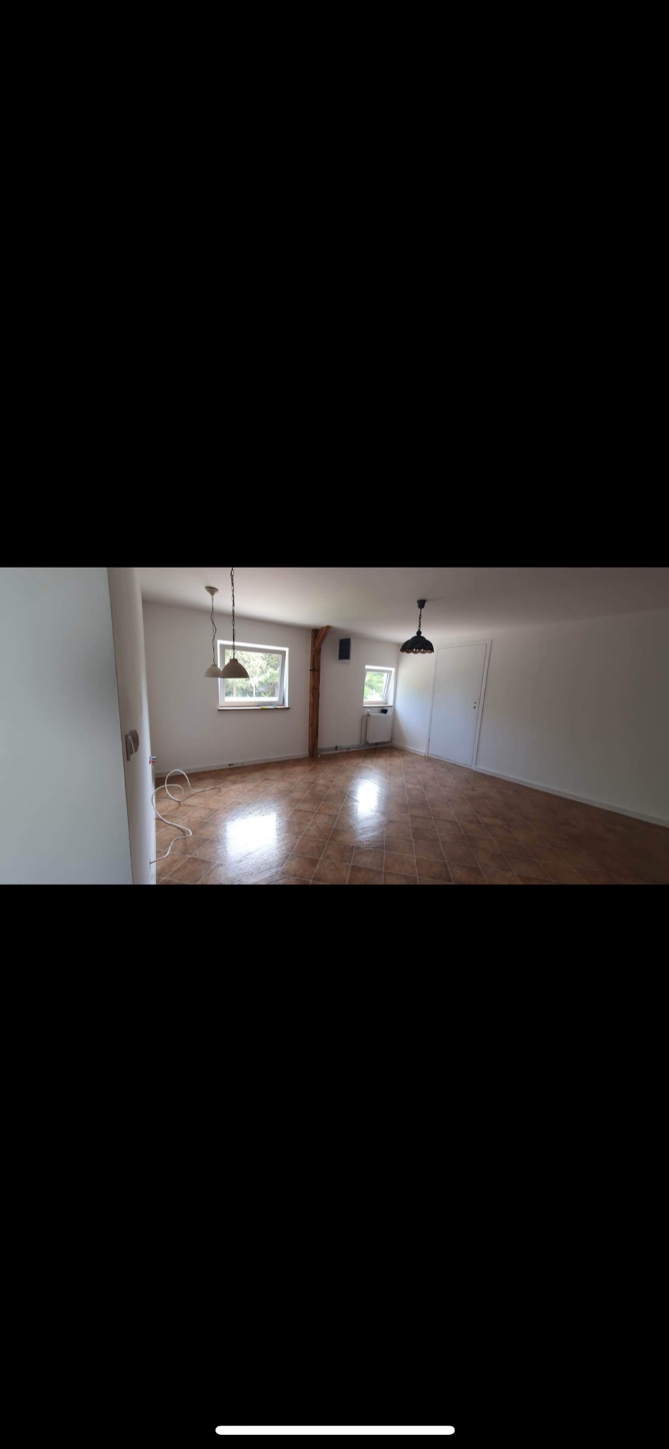 Puste pomieszczenie z brązową terakotą na podłodze, białymi ścianami, dwoma oknami i trzema lampami wiszącymi, widoczny drewniany słup podtrzymujący strop.