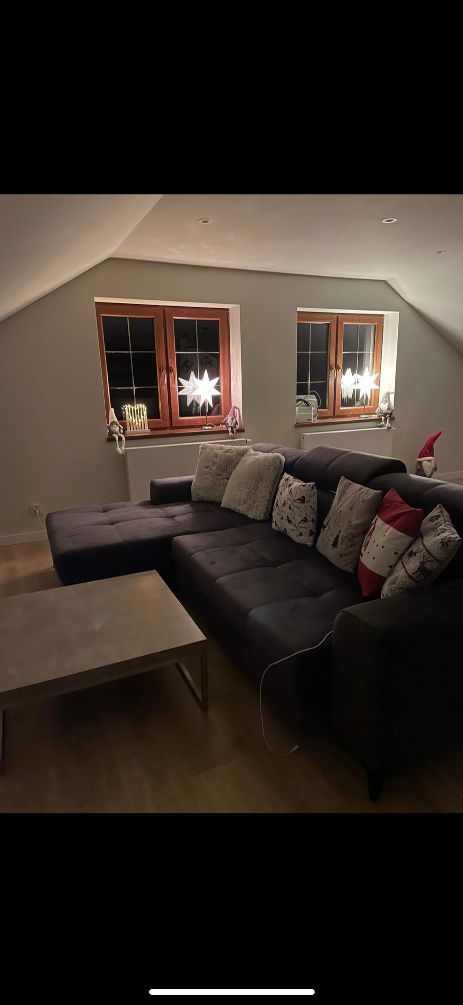 Ciemnoszara sofa narożna z poduszkami dekoracyjnymi w szarym pokoju z dwoma oknami z drewnianymi ramami, w których stoją świecące gwiazdy.