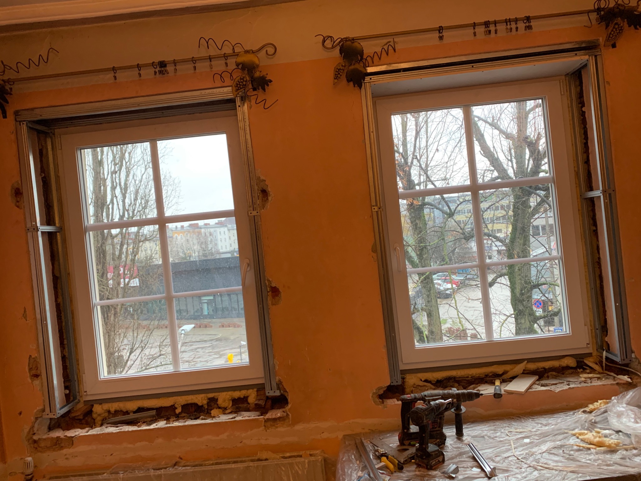 Dwa okna z białymi ramami w trakcie montażu, widoczne metalowe profile mocujące, pianka montażowa i narzędzia na parapecie, ściana w kolorze brzoskwiniowym z dekoracyjnymi elementami nad oknami.