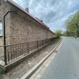 P.H.U. Nikola Atamańczuk-Kolańska - Ceglany budynek z odrapaną elewacją, widoczny zza metalowego ogrodzenia, obok asfaltowa droga z rowerzystą w oddali, pochmurne niebo.