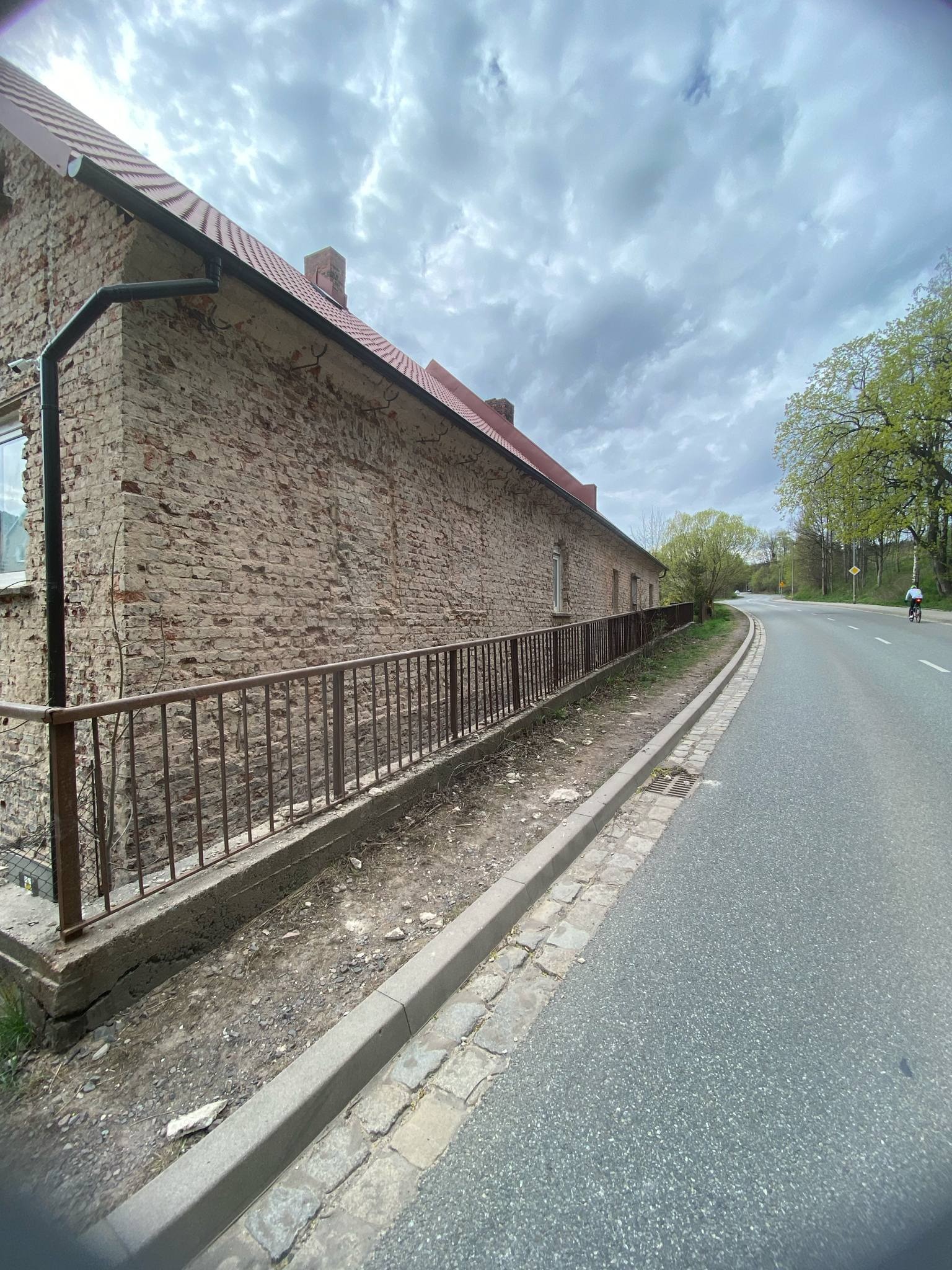 Ceglany budynek z odrapaną elewacją, widoczny zza metalowego ogrodzenia, obok asfaltowa droga z rowerzystą w oddali, pochmurne niebo.