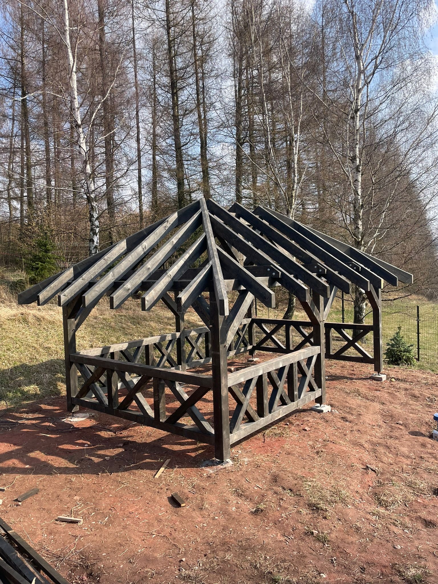 Drewniana altana w trakcie budowy, widoczne elementy konstrukcyjne dachu i ścianek, surowy stan, czerwona ziemia w tle, drzewa bez liści, słoneczny dzień.