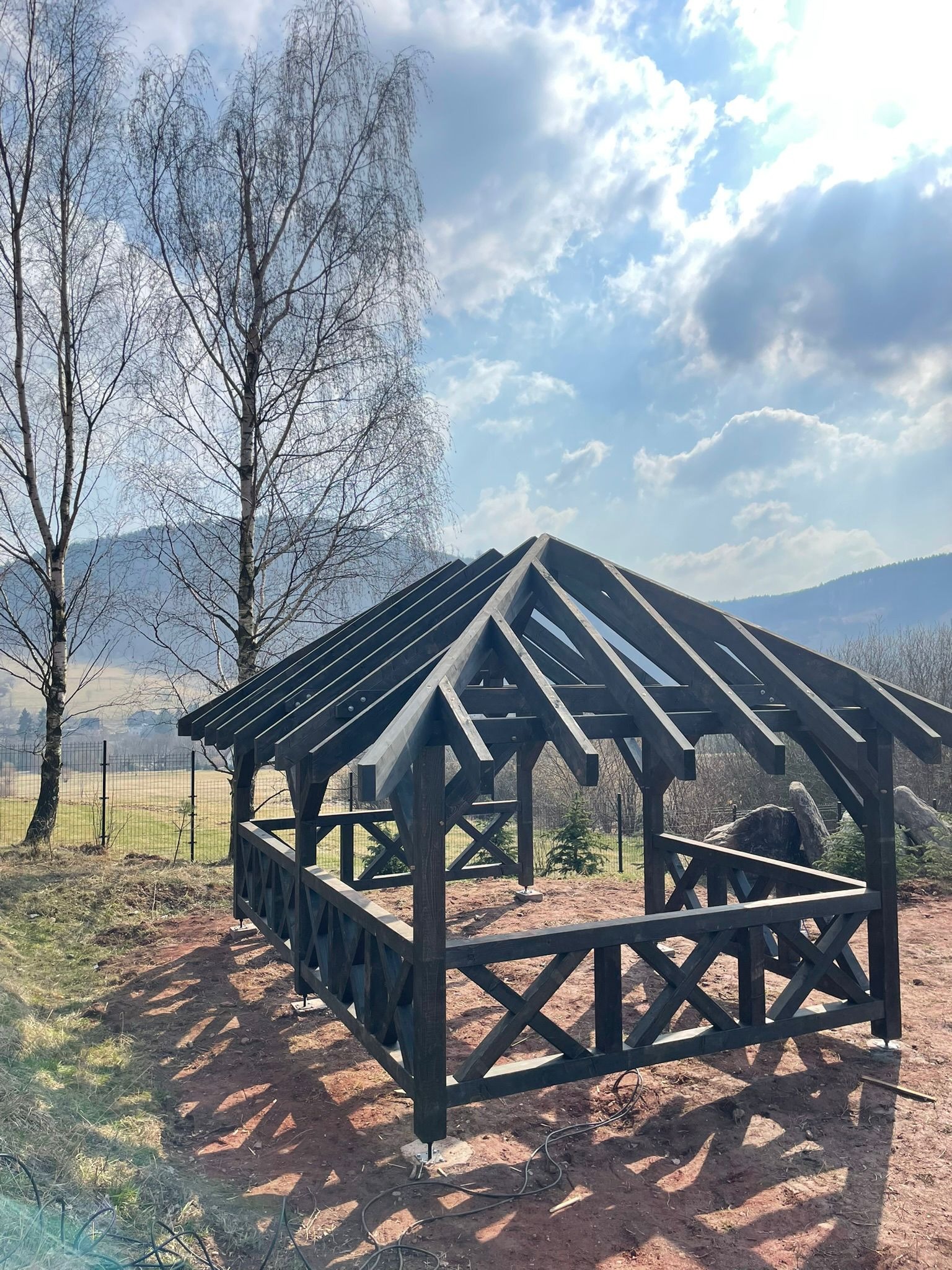 Drewniana altana w trakcie budowy, z widoczną konstrukcją dachu i balustradami, na tle górskiego krajobrazu i błękitnego nieba z chmurami.