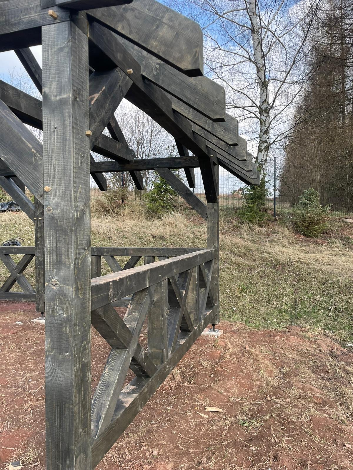 Ciemna, drewniana konstrukcja altany w ogrodzie, z widocznymi elementami dachu i balustrady. Surowe wykończenie drewna, ziemiste podłoże, w tle drzewa i błękitne niebo.