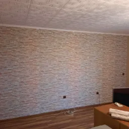 Ściana wykończona tapetą imitującą kamień, z widocznymi rolkami tapety na komodzie i ozdobnym kasetonem na suficie.