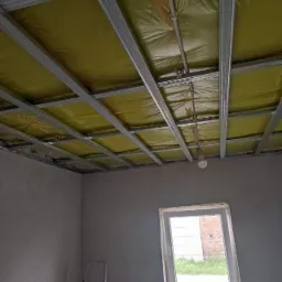 Stelaż sufitowy z izolacją z wełny mineralnej, widok z dołu, surowe ściany w szarym odcieniu, drabina oparta o ścianę, okno z widokiem na ceglany budynek.