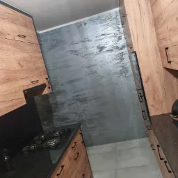 Nowoczesna kuchnia z drewnianymi szafkami z czarnymi uchwytami, czarnym blatem z brokatem i szarą ścianą imitującą beton.