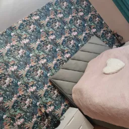 Sypialnia z tapetą w tropikalne wzory, szarą tapicerowaną ścianą za łóżkiem i białą szafką nocną z dwiema szufladami.