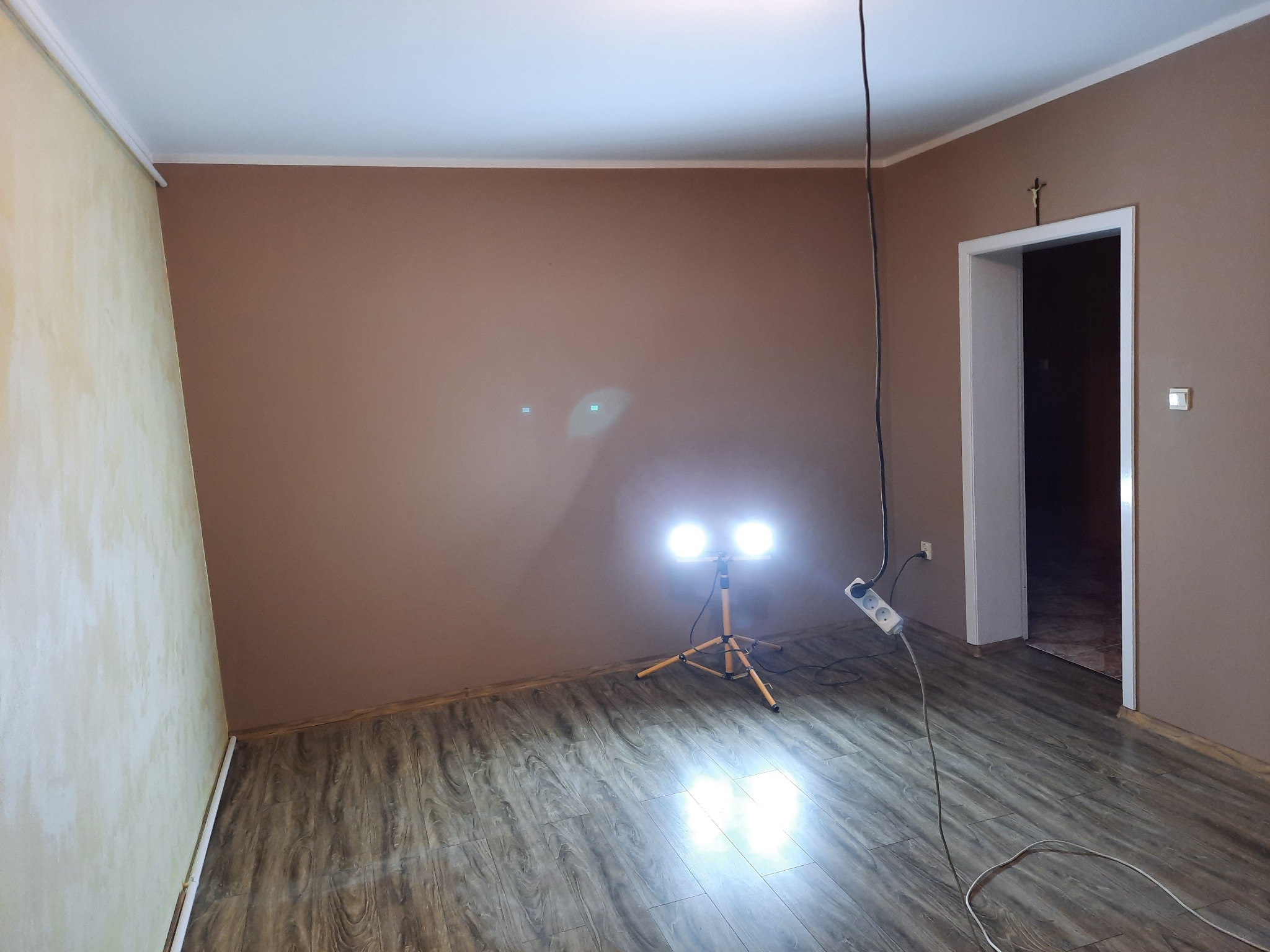 Pokój z brązowymi ścianami w trakcie przygotowań do prac wykończeniowych, oświetlony lampą na statywie, z widocznym gniazdkiem elektrycznym i krzyżem nad drzwiami.
