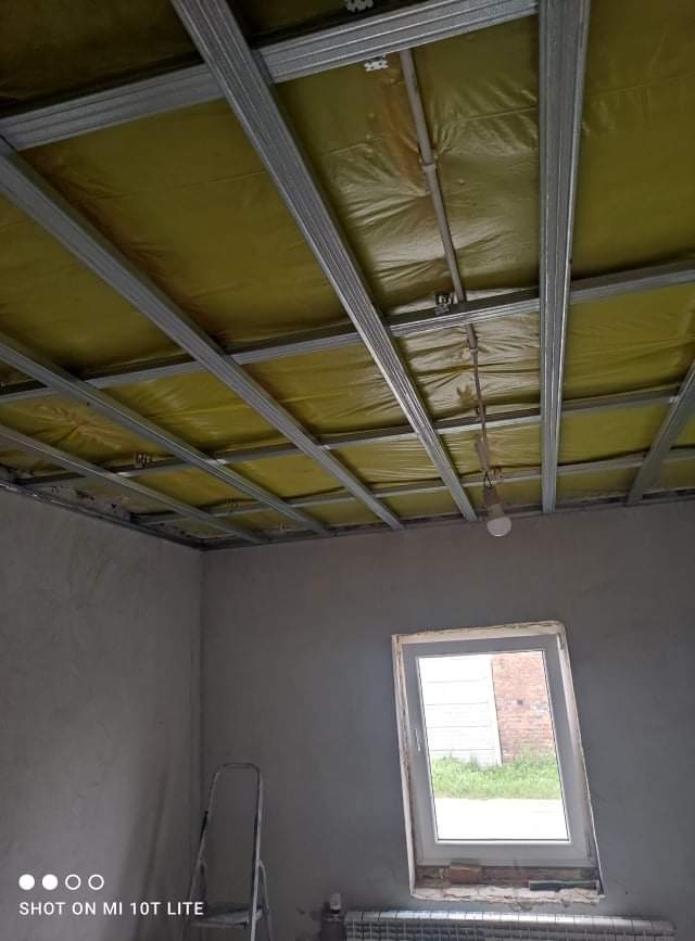 Stelaż sufitowy z izolacją z wełny mineralnej, widok z dołu, surowe ściany w szarym odcieniu, drabina oparta o ścianę, okno z widokiem na ceglany budynek.