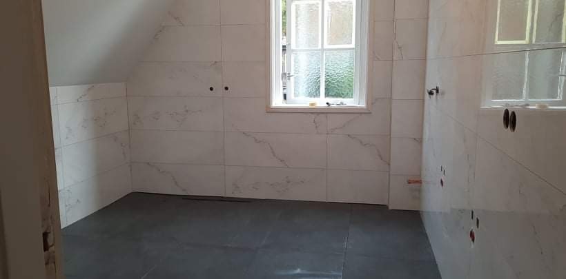 Wnętrze łazienki w trakcie wykańczania: szare płytki podłogowe, białe płytki ścienne imitujące marmur, okno z widokiem na zieleń, widoczne otwory na armaturę.