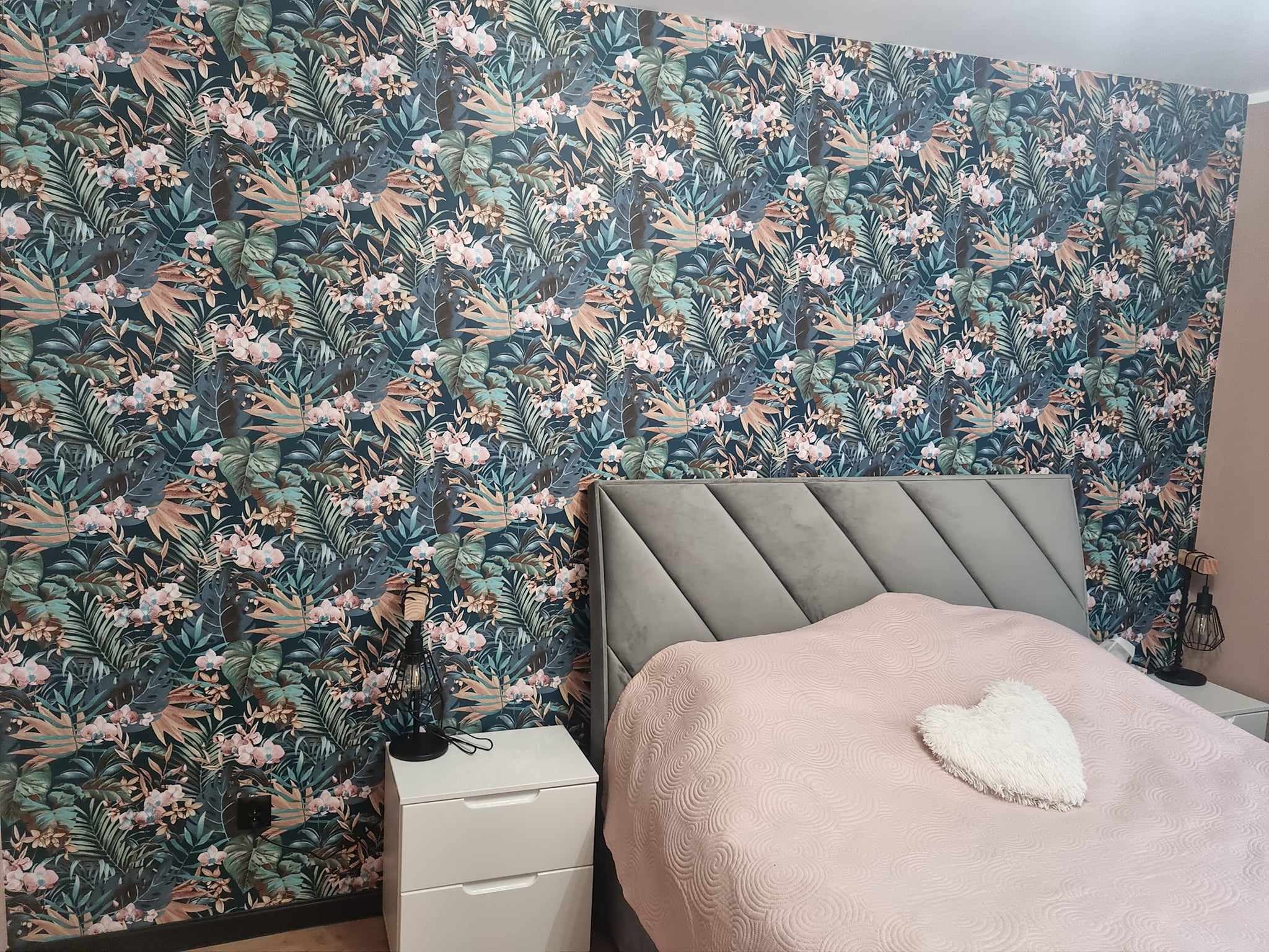 Sypialnia z tapetą w tropikalne liście i kwiaty, szare łóżko z pikowanym zagłówkiem, różowa narzuta z pluszową poduszką w kształcie serca, biała szafka nocna z czarną lampką.