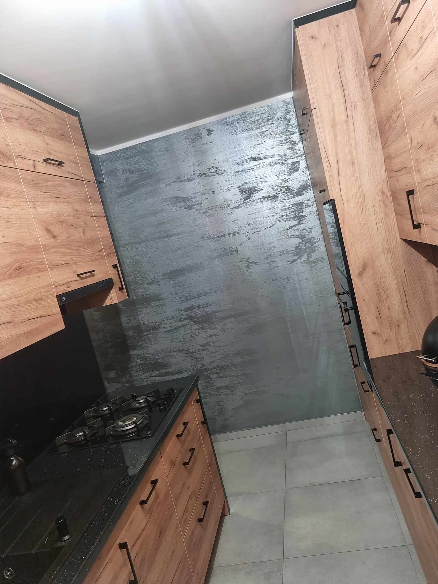 Nowoczesna kuchnia z drewnianymi szafkami z czarnymi uchwytami, czarnym blatem z brokatem i szarą ścianą imitującą beton.