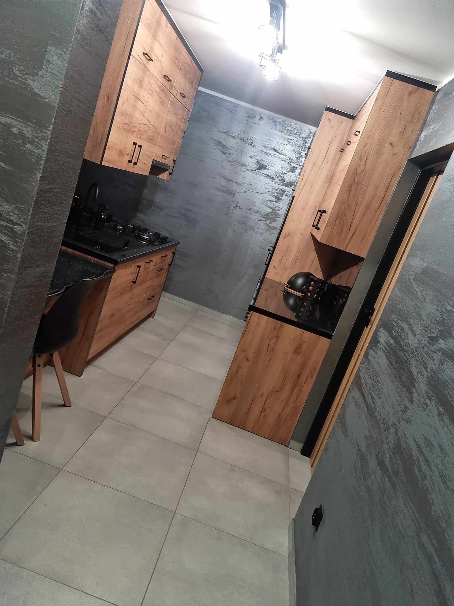 Nowoczesna kuchnia z drewnianymi szafkami i czarnymi blatami, szare ściany z efektem betonu, szara podłoga z płytek, widok z perspektywy podłogi.